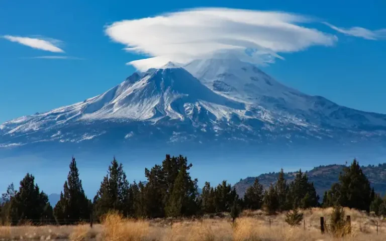 Vista panorámica del Monte Shasta, mostrando la majestuosidad de la montaña rodeada de árboles, nubes flotantes y un cielo despejado, capturando la serenidad y energía espiritual del paisaje.