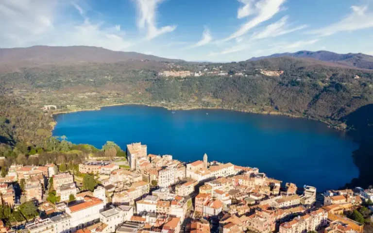 Vista panorámica del lago de Nemi con la ciudad a la orilla, rodeado de bosques verdes bajo un cielo azul despejado.