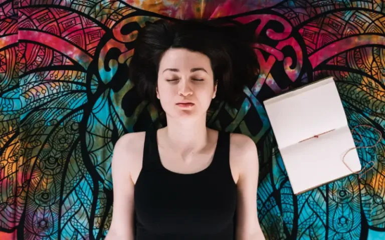 Vista aérea de una mujer con los ojos cerrados, acostada cerca de un libro abierto sobre una manta, acompañada de un cuaderno, simbolizando la introspección profunda y la meditación espiritual.